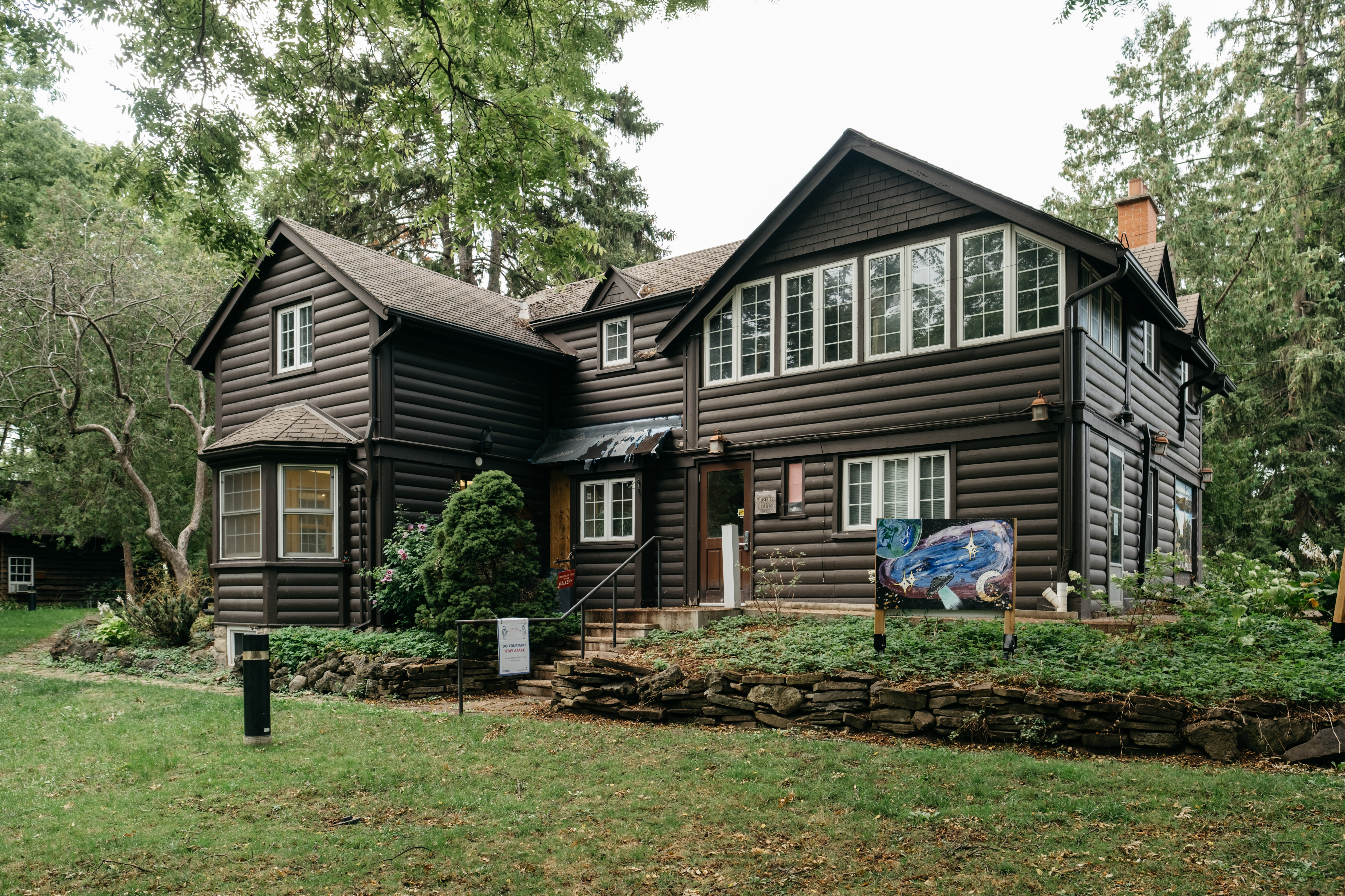 A two-storey log-clad house with gabled roofs set within a woodsy landscape of mature trees. 